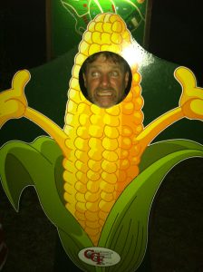 Chris is smiling with his face visible from the cutout on a large painting of an ear of corn.